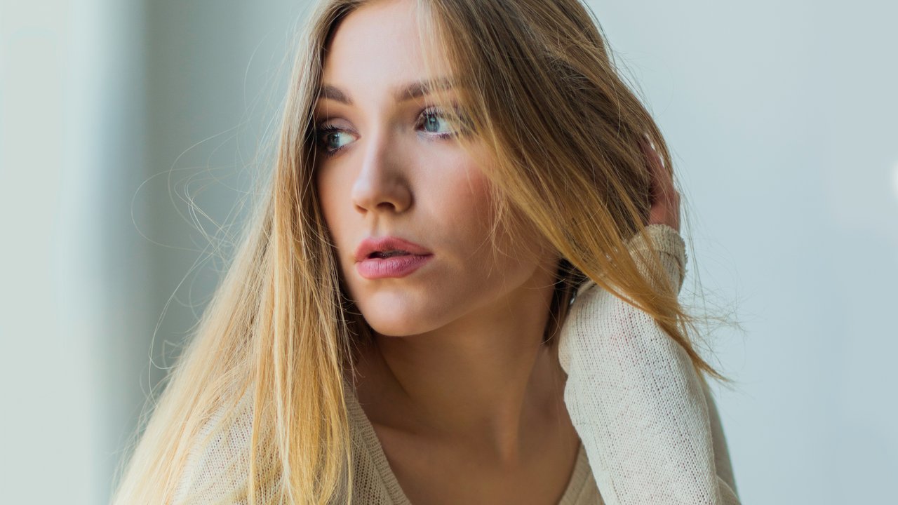 Young blonde woman sitting next to window in sweater, looking emotive and thoughtful