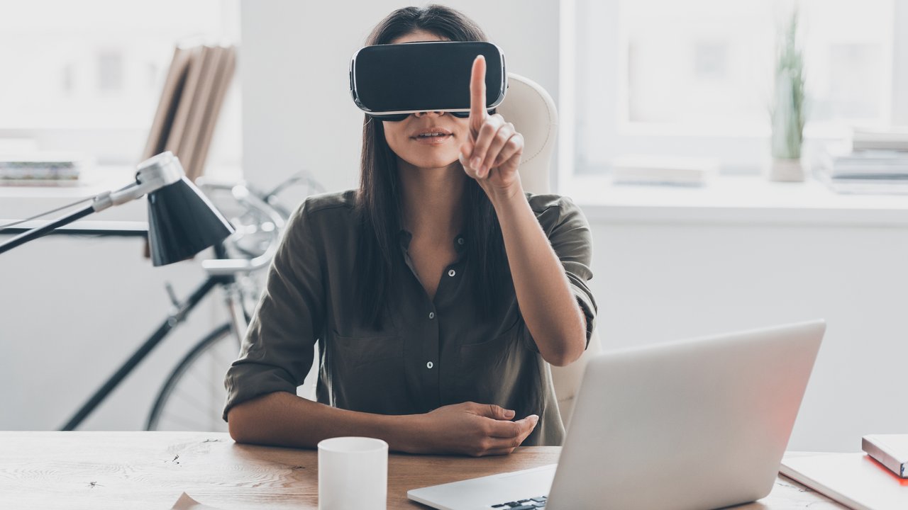 Confident young woman in virtual reality headset pointing in the air while sitting at her working place in office