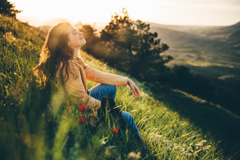 Eine Frau mit langen, lockeren Locken sitzt auf einer grünen Graswiese mit Blumen vor einer hügeligen Landschaft im Sonnenlicht.