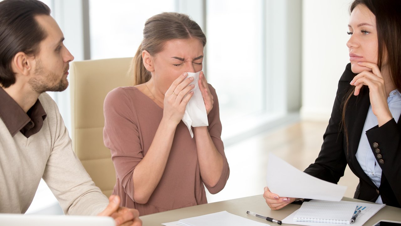 Sick young attractive woman with handkerchief has sneezing attack, blowing nose while working with colleagues on meeting, caught cold, flu symptom, weakened immune system due to stress or overwork