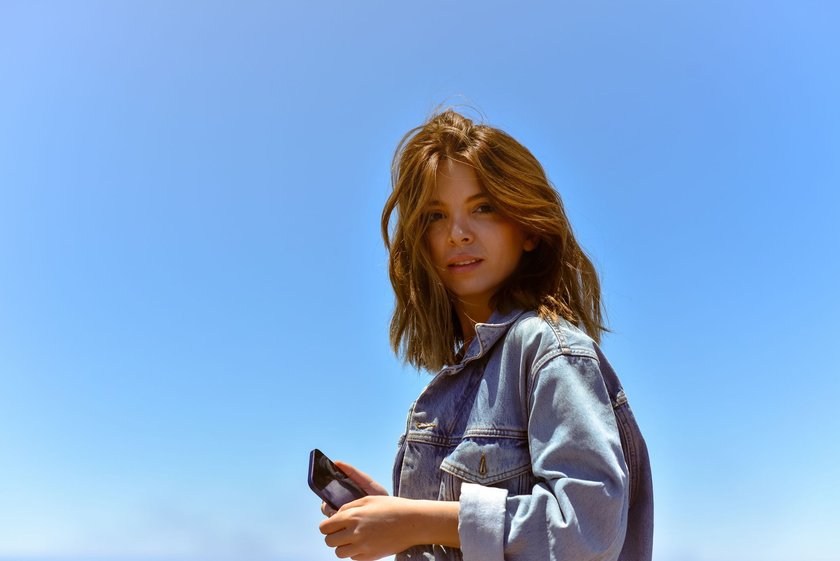 Eine&#x20;Frau&#x20;mit&#x20;braunen&#x20;Haaren&#x20;tr&#x00E4;gt&#x20;eine&#x20;Jeansjacke&#x20;und&#x20;h&#x00E4;lt&#x20;ein&#x20;Smartphone