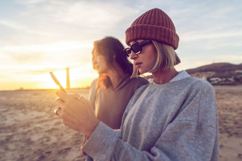zwei Frauen am Strand, die eine schaut auf ihr Handy