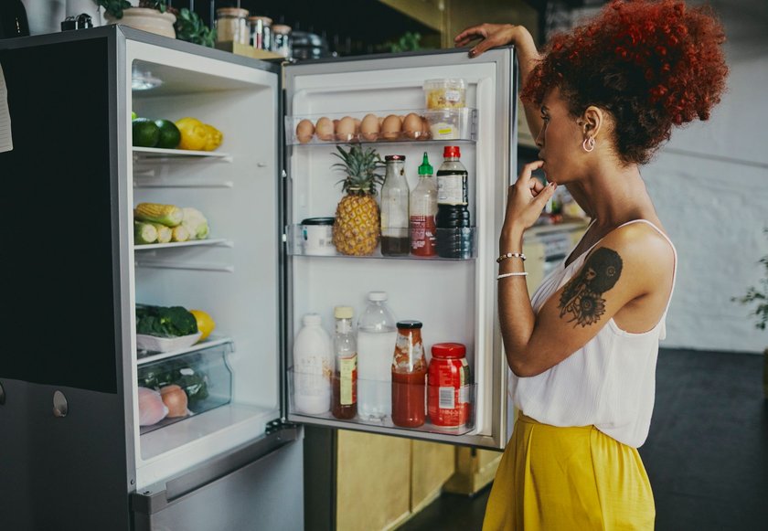 Woman stands in front of an open refrigerator stocked with healthy fruits and vegetables, stock photo