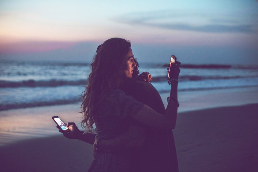 Paar, das sich am Strand umarmt und aufs Handy schaut