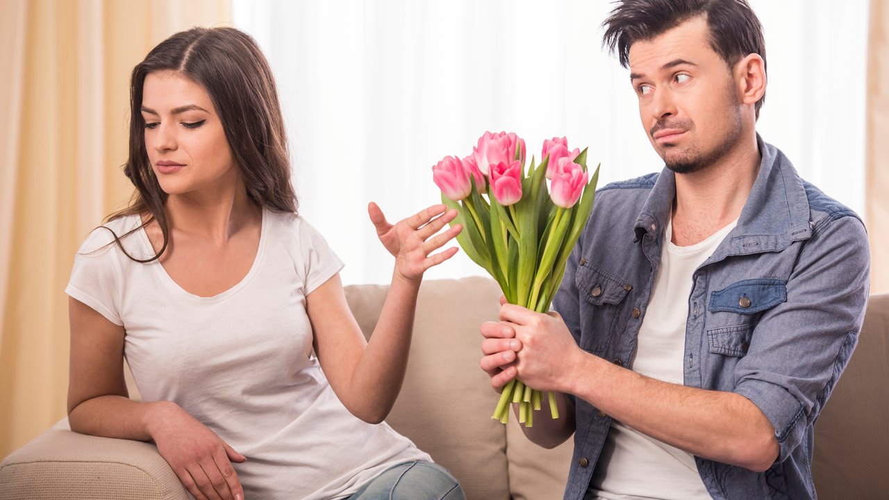 Young man is offering a bunch of flowers to his angry girlfriend at home.