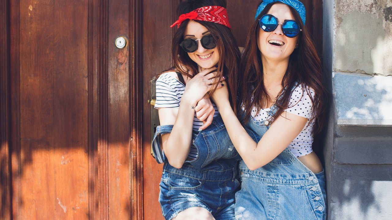 Beautiful young brunette twins sisters, in stylish sunglasses, hugging and laughing. Having fun time together. Wearing denim overalls bright bandanas, posing in front of old doors. Outdoors. Summer day. Copy space.