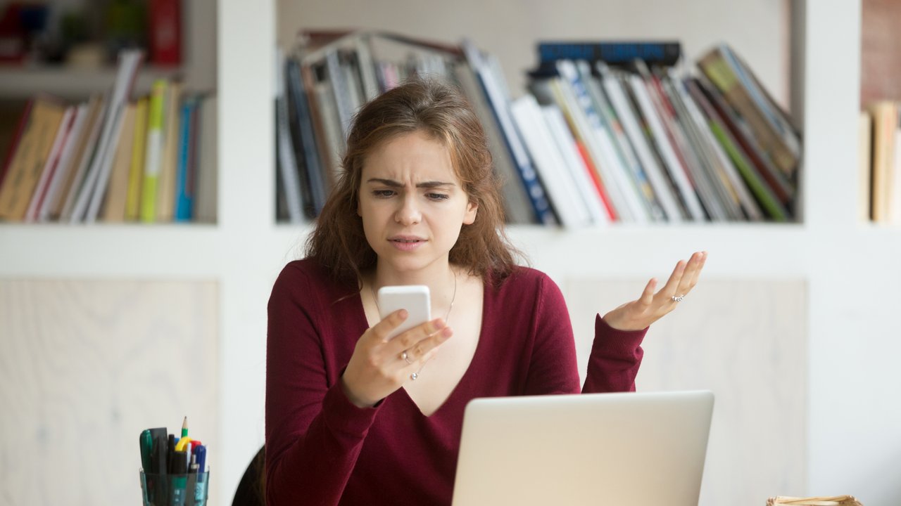 Frustrated female entrepreneur looks at cellphone and shrugs. Young businesswoman unhappy with cellular provider service, smartphone battery died. Freelance business owner received high phone bill.