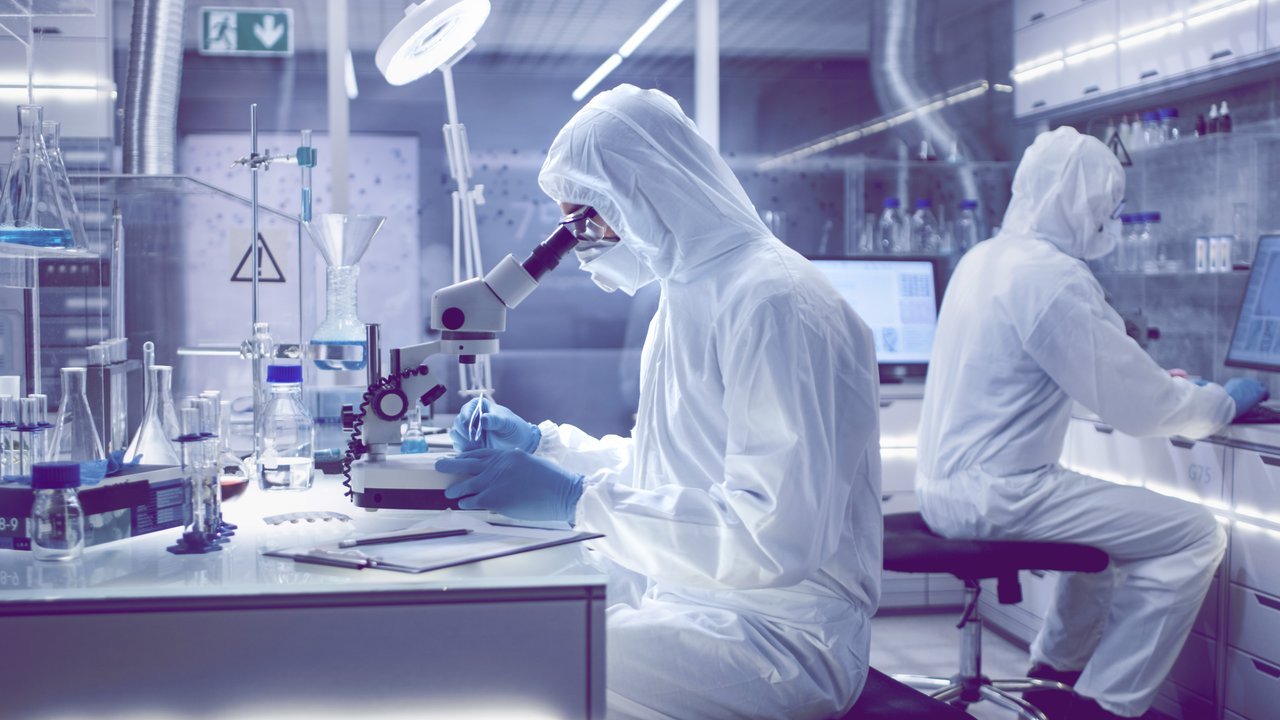 In a Secure High Level Laboratory Scientists in a Coverall Conducting a Research. Biologist Adjusts Samples in a  Petri Dish with Pincers and Examines Them Under Microscope and His Colleague Analyzes Results on a Computer.