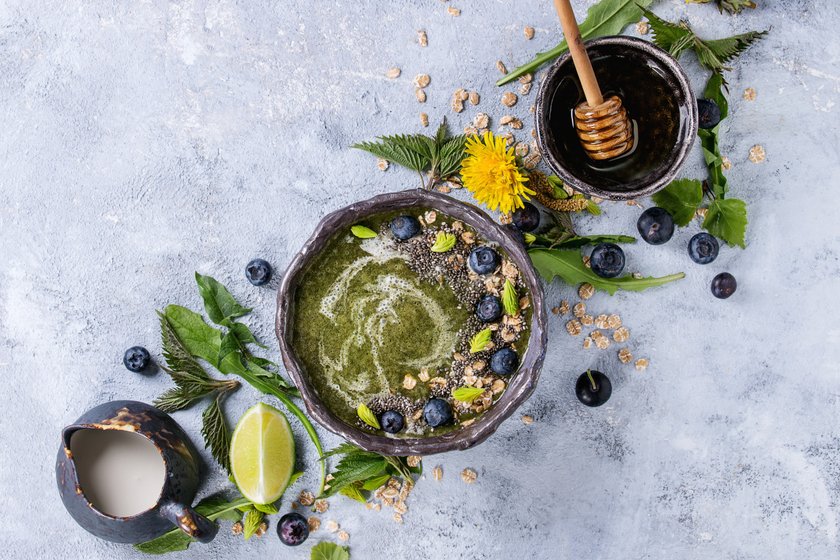 Spring green nettle and dandelion smoothie bowl served with lime, yellow flowers, young leaves, oat flakes, chia seeds, blueberries, cream and honey over gray blue texture background. Top view, space. (Photo by: Natasha Breen/REDA/Universal Images Group via Getty Images)