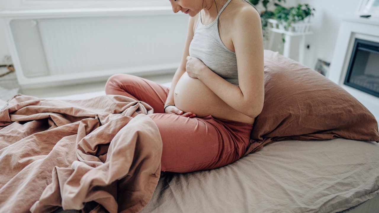 Photo of a young expectant mother stroking her baby bump and enjoying her morning in the bedroom of her apartment; the daily routine of a pregnant woman.