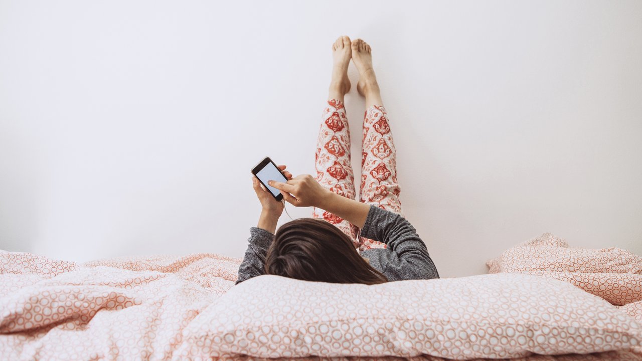 The girl lies in bed and listens to music or a podcast. The girl uses her cell phone.