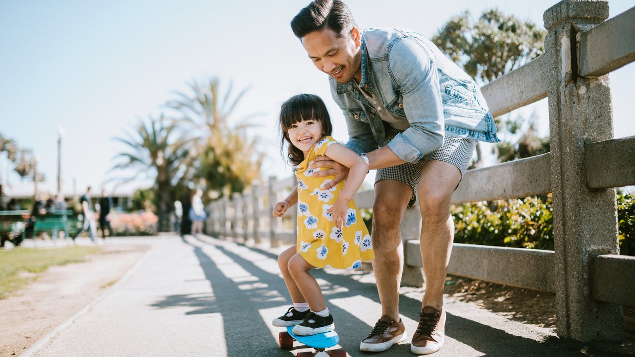 A dad helps his little girl go skateboarding, holding her waist for support.  Shot in Los Angeles, California by the Santa Monica Pier.