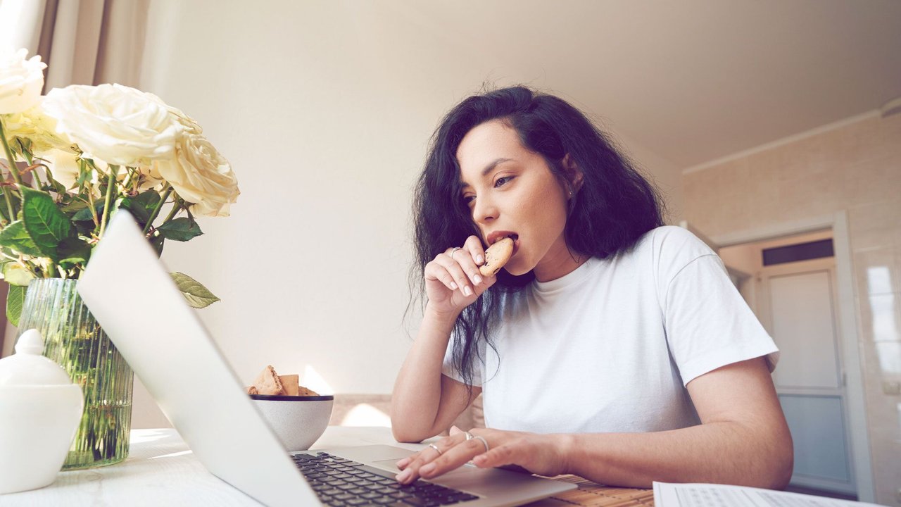 Serious focused young female eating cookie at table and typing on laptop