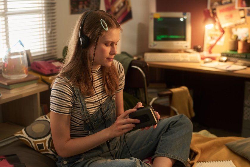 Young teenager listening to music with headphones in a retro-themed room with vintage decor and ambience, holding electronic device while sitting on chair
