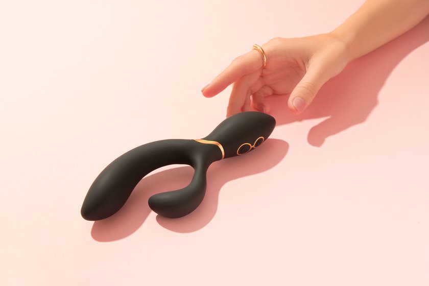 Female hand with pink polish nails is touching a black vibrator which lies on the peach pink background. Sex toys for adults