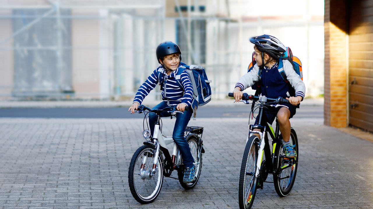 Two school kid boys in safety helmet riding with bike in the city with backpacks. Happy children in colorful clothes biking on bicycles on way to school. Safe way for kids outdoors to school.