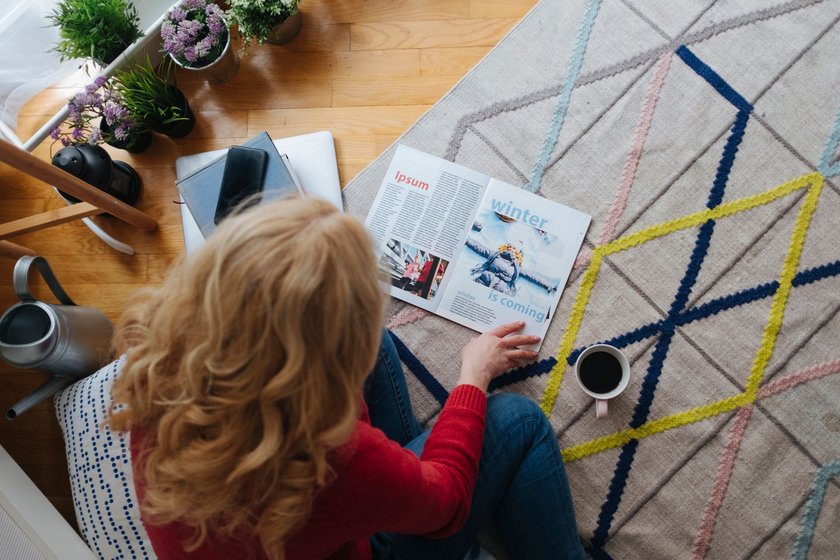 Mature woman sitting on the floor in her living room, drinking coffee and reading magazine.