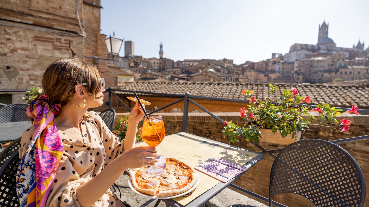 Young woman having lunch with pizza and wine at outdoor restaurant with beautiful view on the old town of Siena. Concept of italian cuisine and traveling Tuscany region of Italy
