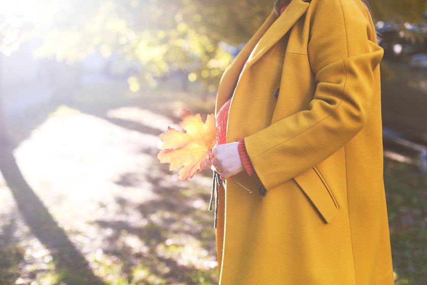 Schwangere Frau mit gelben Mantel steht in der Sonne und halt ein Blatt in der Hand