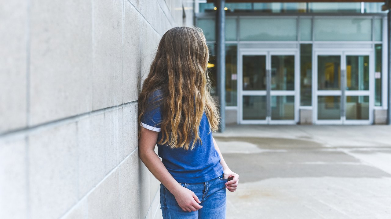 The image displays a teenage girl leaning against a school wall all alone as she glances over her shoulder.