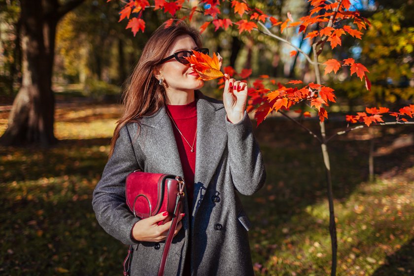 Happy stylish woman wears trendy autumn outfit. Woman wearing grey fall coat, holding burgundy leather purse outdoors. Fall fashion