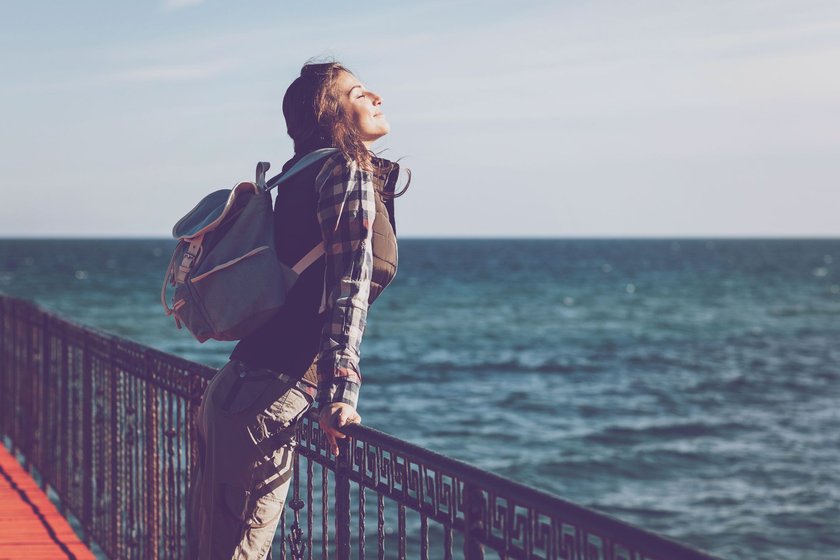Frau, die mit Rucksack an einem Zaun steht und aufs Wasser schaut