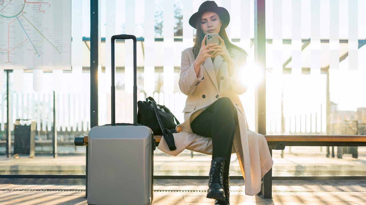Tourist girl with suitcase sits at public transport stop and looks in smartphone. Girl waiting for the bus