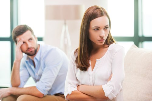 Depressed young woman keeping arms crossed and looking away while man sitting behind her on the couch Depressed young woman keeping arms crossed and looking away while man sitting behind her on the couch