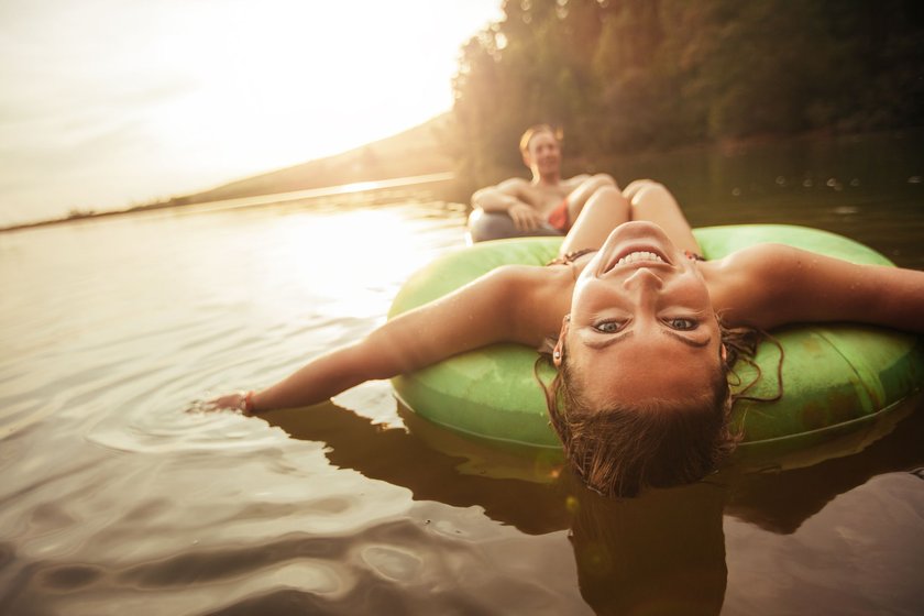 Porträt einer glücklichen jungen Frau, die in einem Schwimmreifen auf dem See treibt, während ihr Freund im Hintergrund ist.