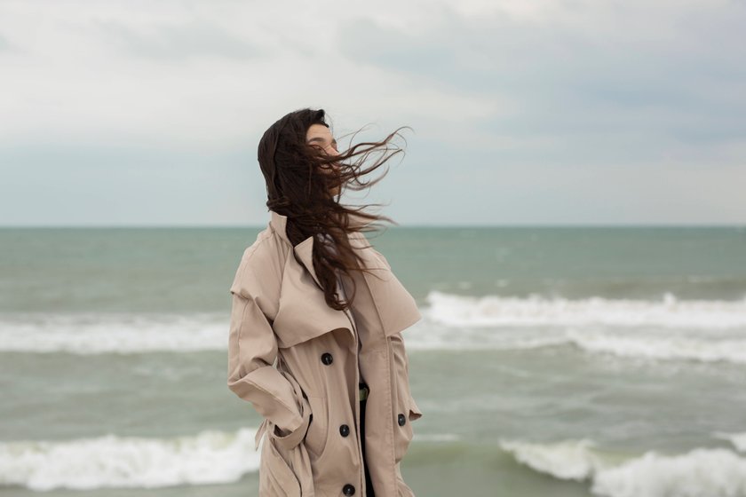 Eine Frau mit schwarzem, wehendem Haar in einem Mantel steht an einem Strand bei windigem Wetter