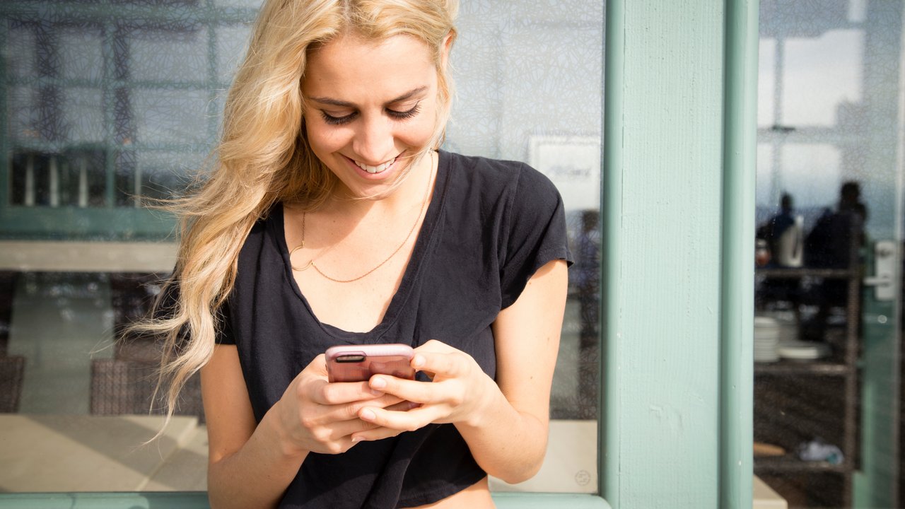 Close up shot of a pretty young woman texts on her mobile phone. She is smiling and happy, She looks like a California Beach girl. She is outside on a summer day on a boardwalk.