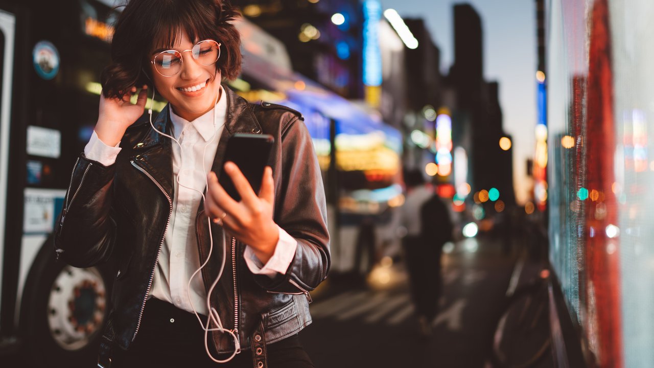 Cheerful pretty young woman in cool eyeglasses and trendy wear walking on metropolis street with night lights enjoying audio songs from playlist in earphones connected to smartphone device