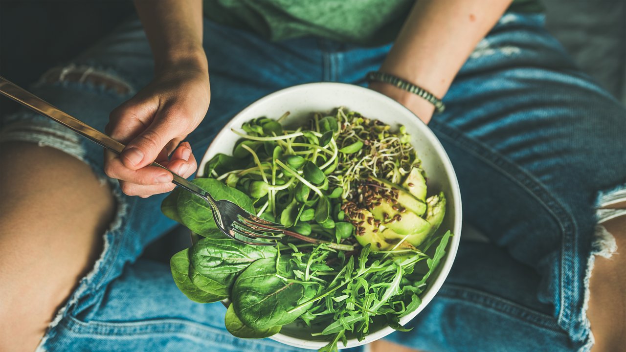 Green vegan breakfast meal in bowl with spinach, arugula, avocado, seeds and sprouts. Girl in jeans holding fork with knees and hands visible, top view. Clean eating, dieting, dieting food concept
