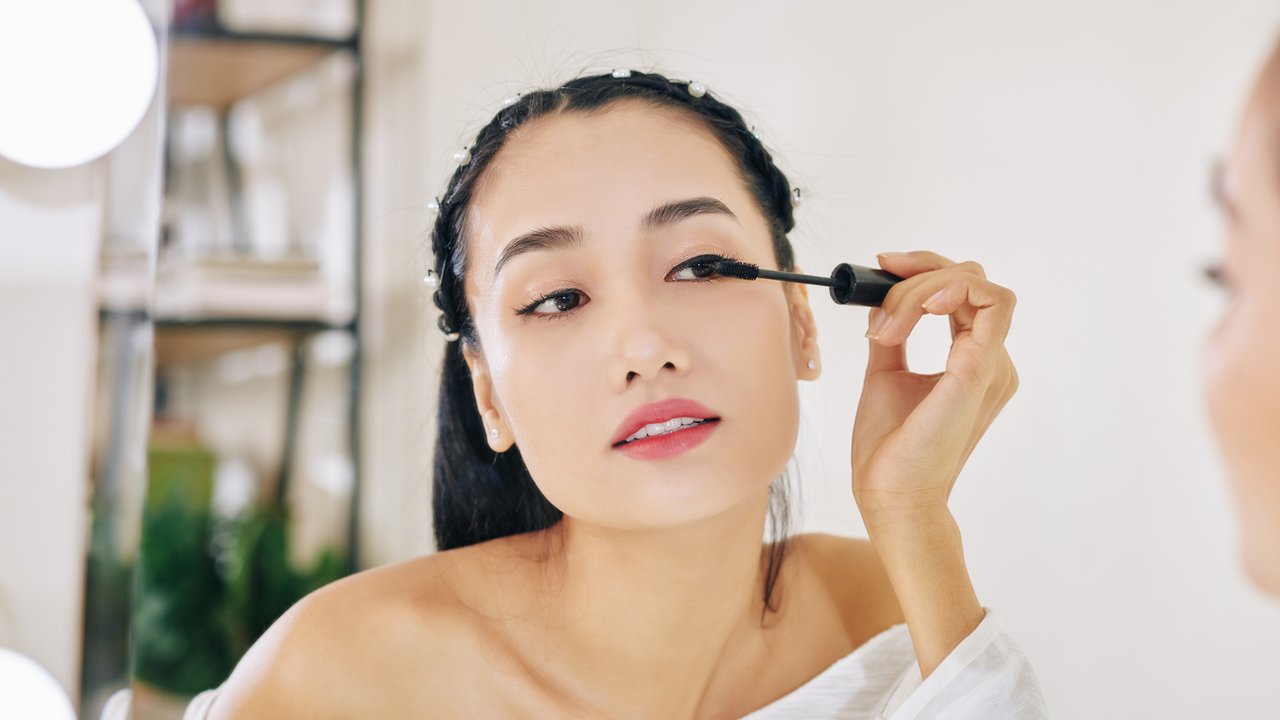 Pretty young Asian woman looking at mirror and applying second coat of black mascara
