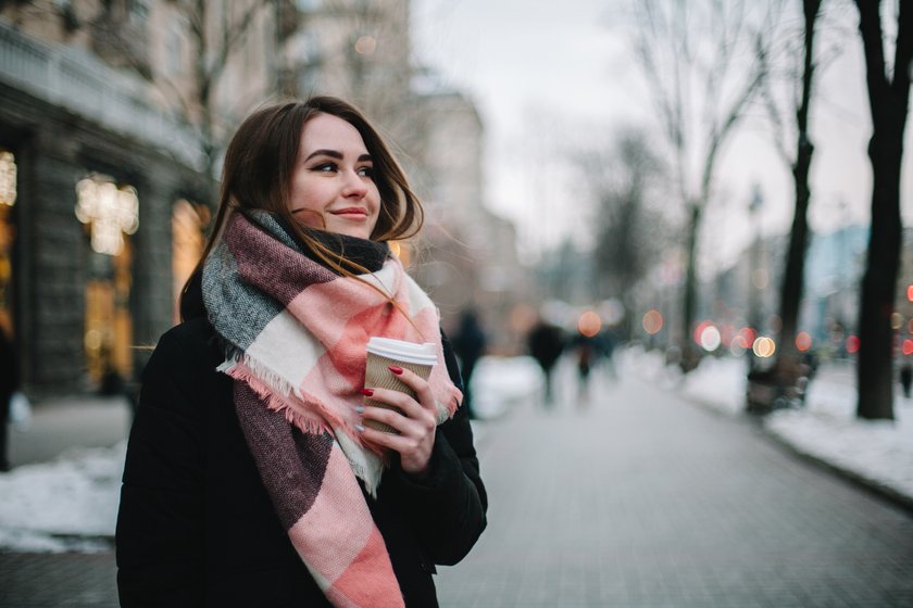 Happy young woman with disposable cup wearing warm clothing while standing on city street during winter