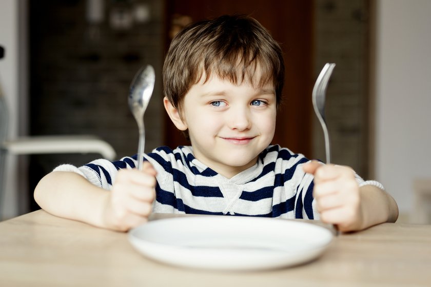 Junge sitzt mit Besteck in der Hand am Tisch und wartet auf Essen